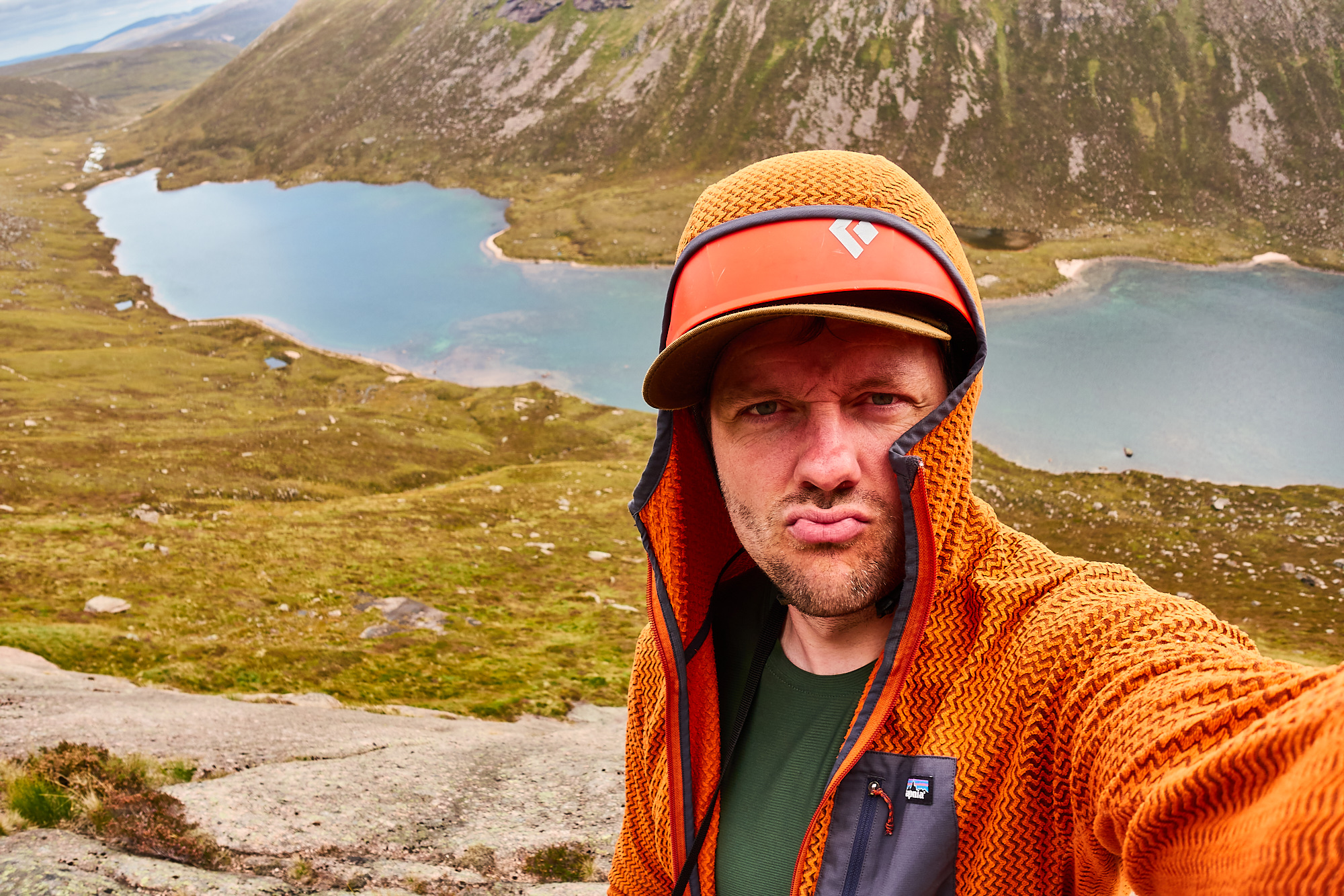 A climber dressed in an orange fleece with a red helmet stands pouting at the camera as he takes a picture of himself, with a blue loch behind and green heather-clad slopes