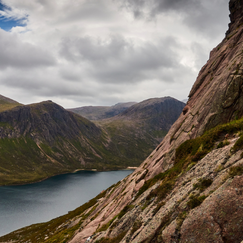 A view of sweeping pale pink granite rock slabs with vertical cracks beneath a cloudy blue sky. A dark blue loch in the background surrounded by grey craggy cliffs and steep slopes