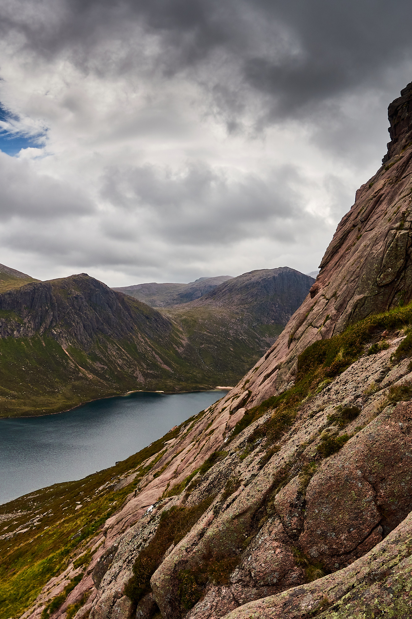 A view of sweeping pale pink granite rock slabs with vertical cracks beneath a cloudy blue sky. A dark blue loch in the background surrounded by grey craggy cliffs and steep slopes