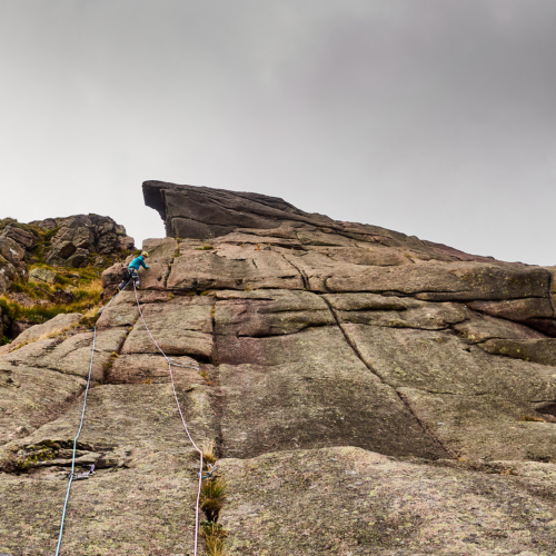 A climber dressed in bright blue with two ropes trailing behind climbs a grey and pink granite slab covered in vertical cracks. A giant overhang of rock protrudes above the climber and the sky is a moody cloudy grey