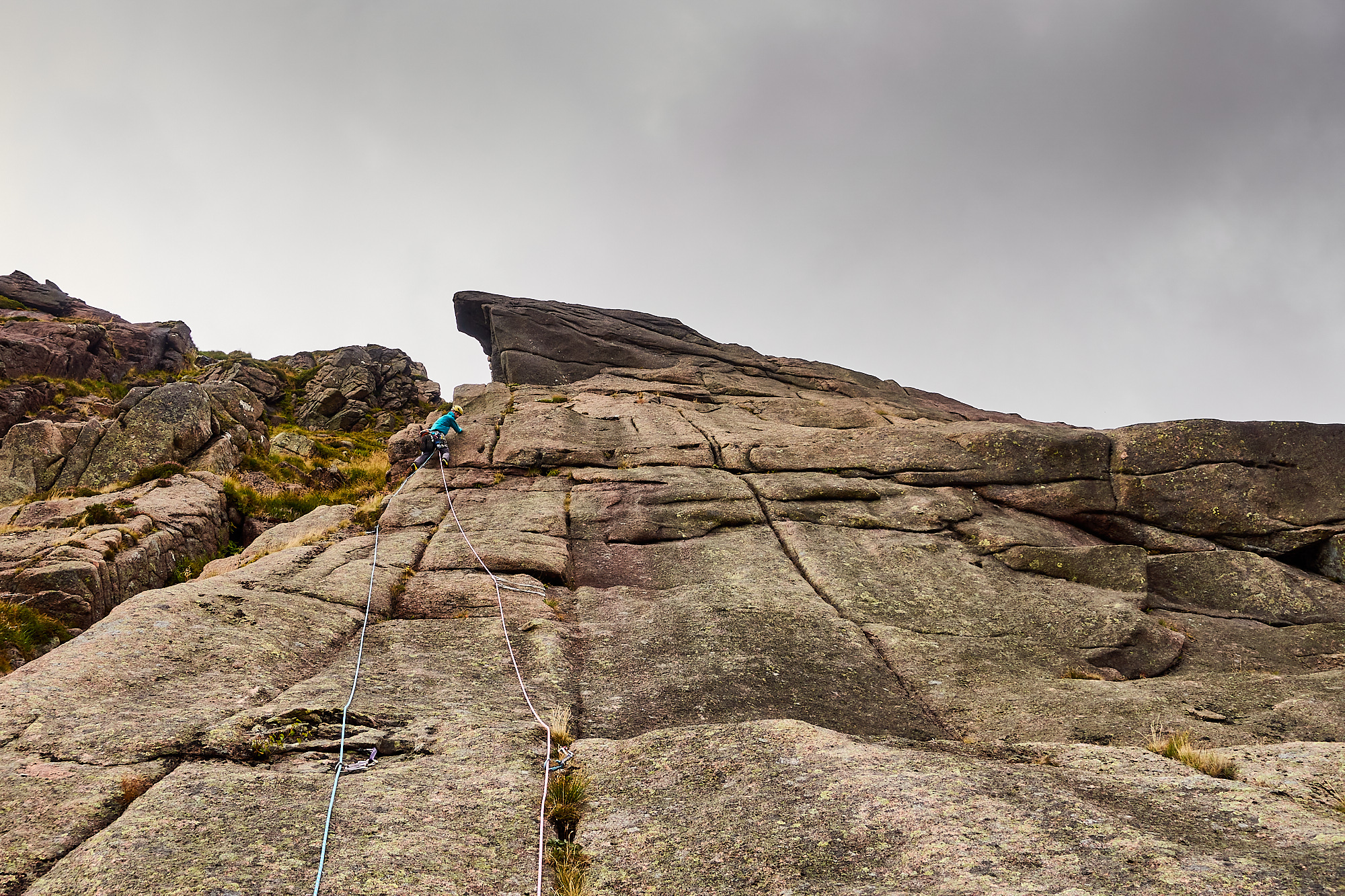 A climber dressed in bright blue with two ropes trailing behind climbs a grey and pink granite slab covered in vertical cracks. A giant overhang of rock protrudes above the climber and the sky is a moody cloudy grey