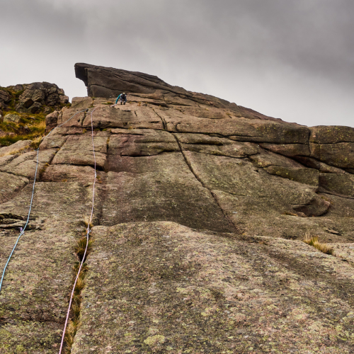 A climber dressed in bright blue with two ropes trailing behind climbs a grey and pink granite slab covered in vertical cracks. A giant overhang of rock protrudes above the climber and the sky is a moody cloudy grey