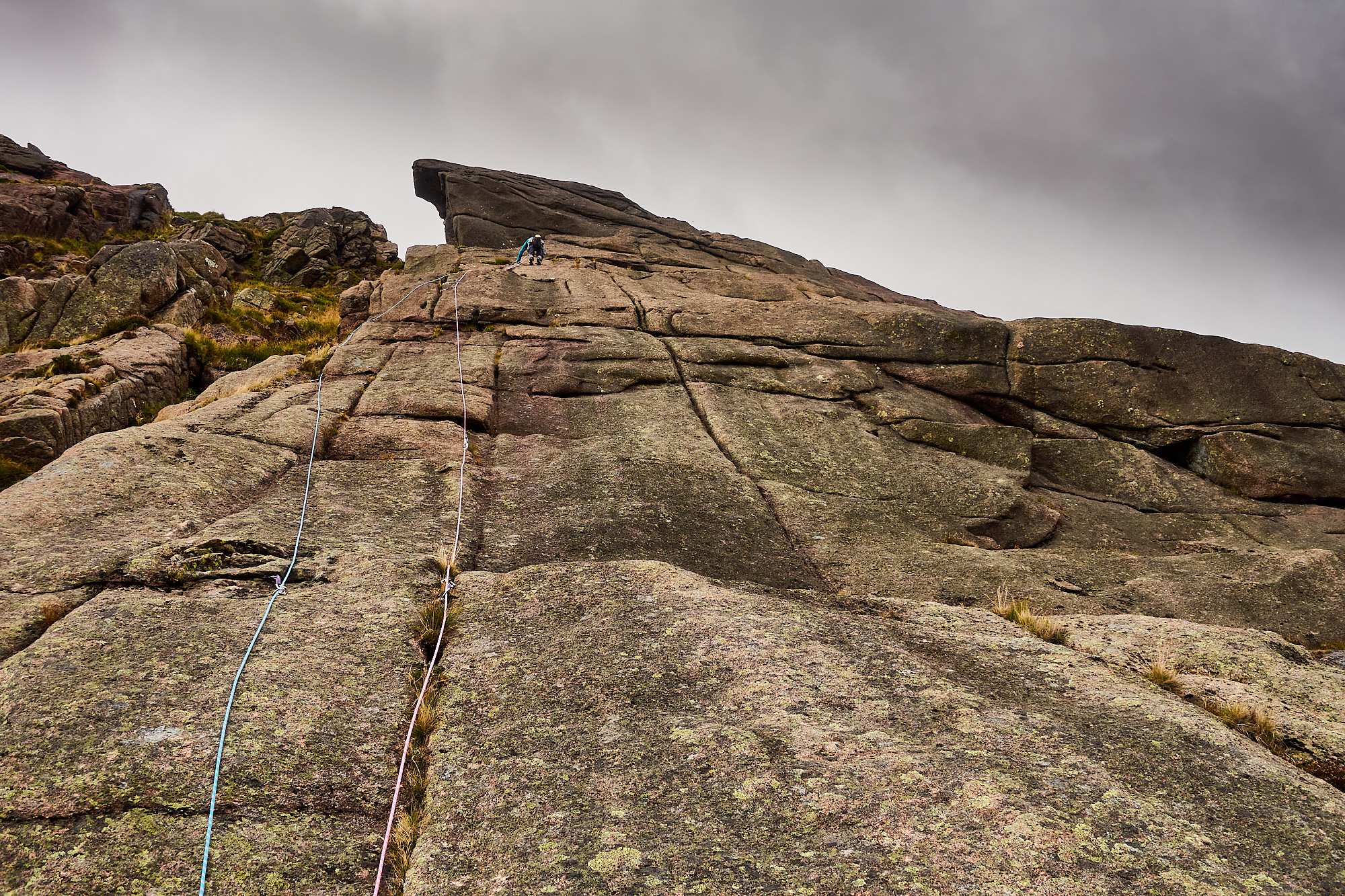 A climber dressed in bright blue with two ropes trailing behind climbs a grey and pink granite slab covered in vertical cracks. A giant overhang of rock protrudes above the climber and the sky is a moody cloudy grey
