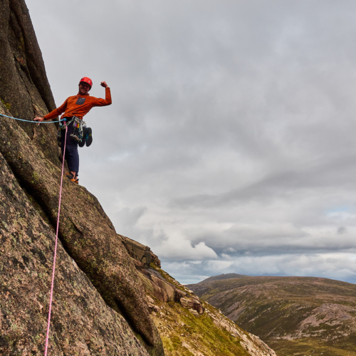 A climber dressed in an orange fleece with a red helmet stands beneath a bulge of rock on a lichen-covered slab, flexing their biceps. Rounded mountains and cloudy skies are behind.