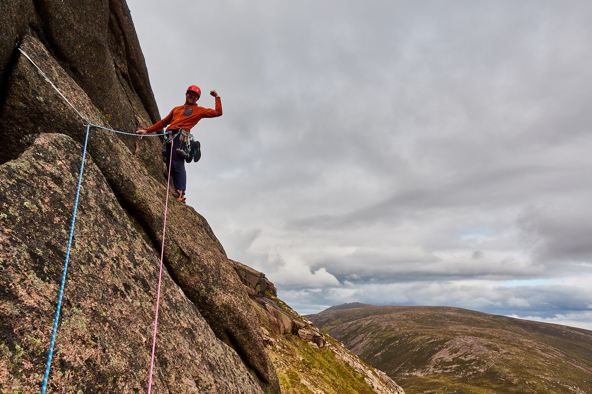 A climber dressed in an orange fleece with a red helmet stands beneath a bulge of rock on a lichen-covered slab, flexing their biceps. Rounded mountains and cloudy skies are behind.