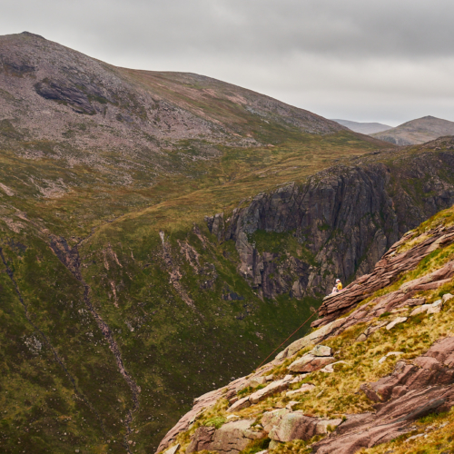 A climbers sits on top of a pink slab of rock above a huge drop with more cliffs and mountains across the valley behind them