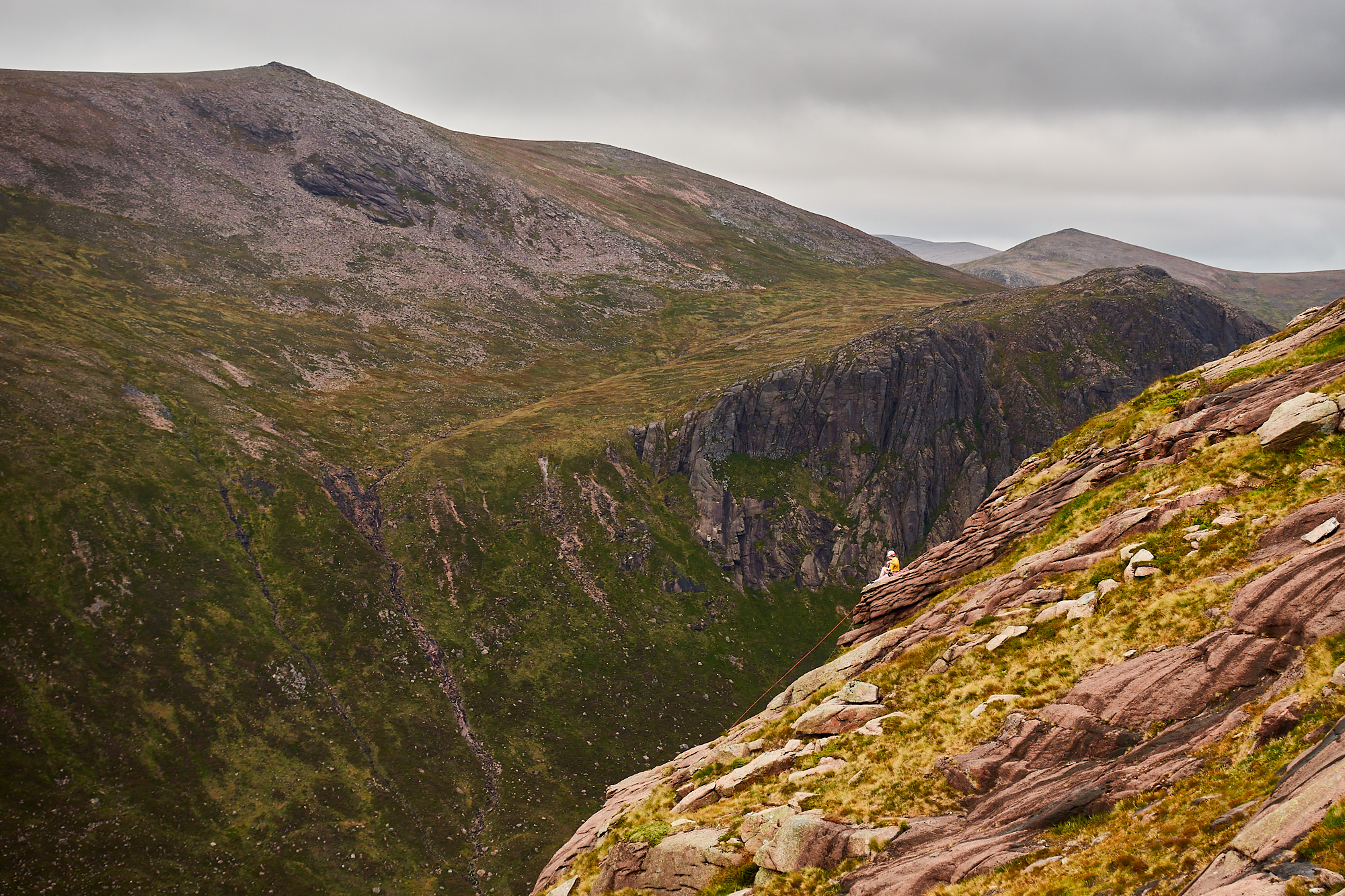 A climbers sits on top of a pink slab of rock above a huge drop with more cliffs and mountains across the valley behind them