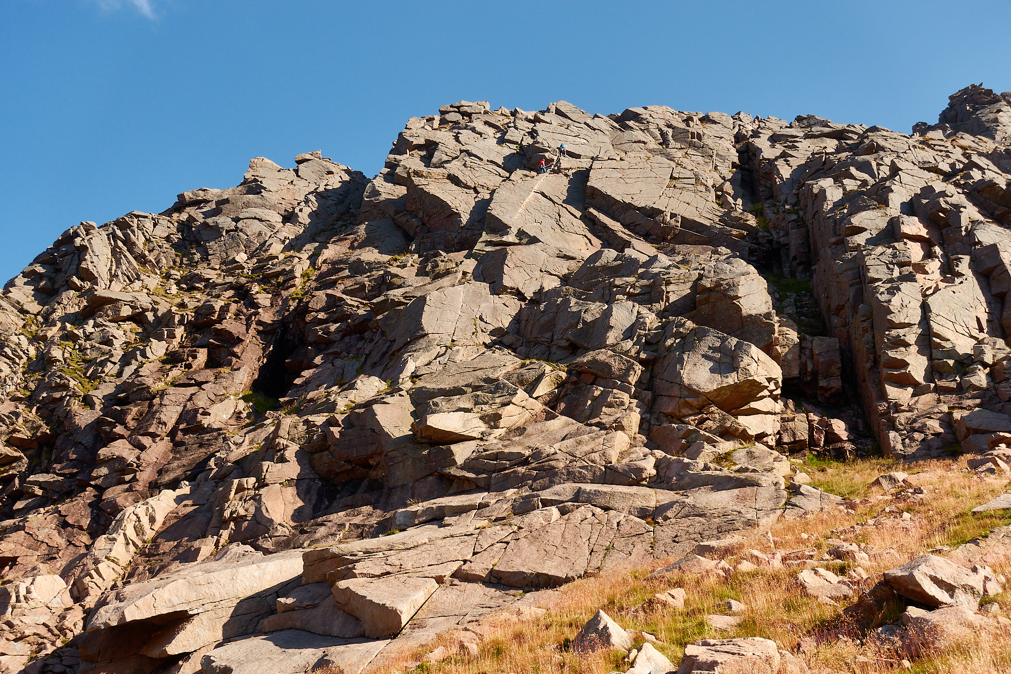 Two climbers near the top of a blocky grey and pink granite cliff, above a striking diagonal crack in a clean slab. Blue sky above and the sun is shining on the cliff.